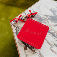 A red square tin with the brand name 'Essentials' embossed on the lid, tied with a red ribbon, resting on a white and gray marble-patterned table next to a green outdoor surface.