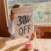 A person sitting at a wooden table by a large window holds up a cardboard sign reading '30% OFF'. The scene includes a red-and-white striped mug, a lit candle, and an outdoor landscape with mountains visible in the background.