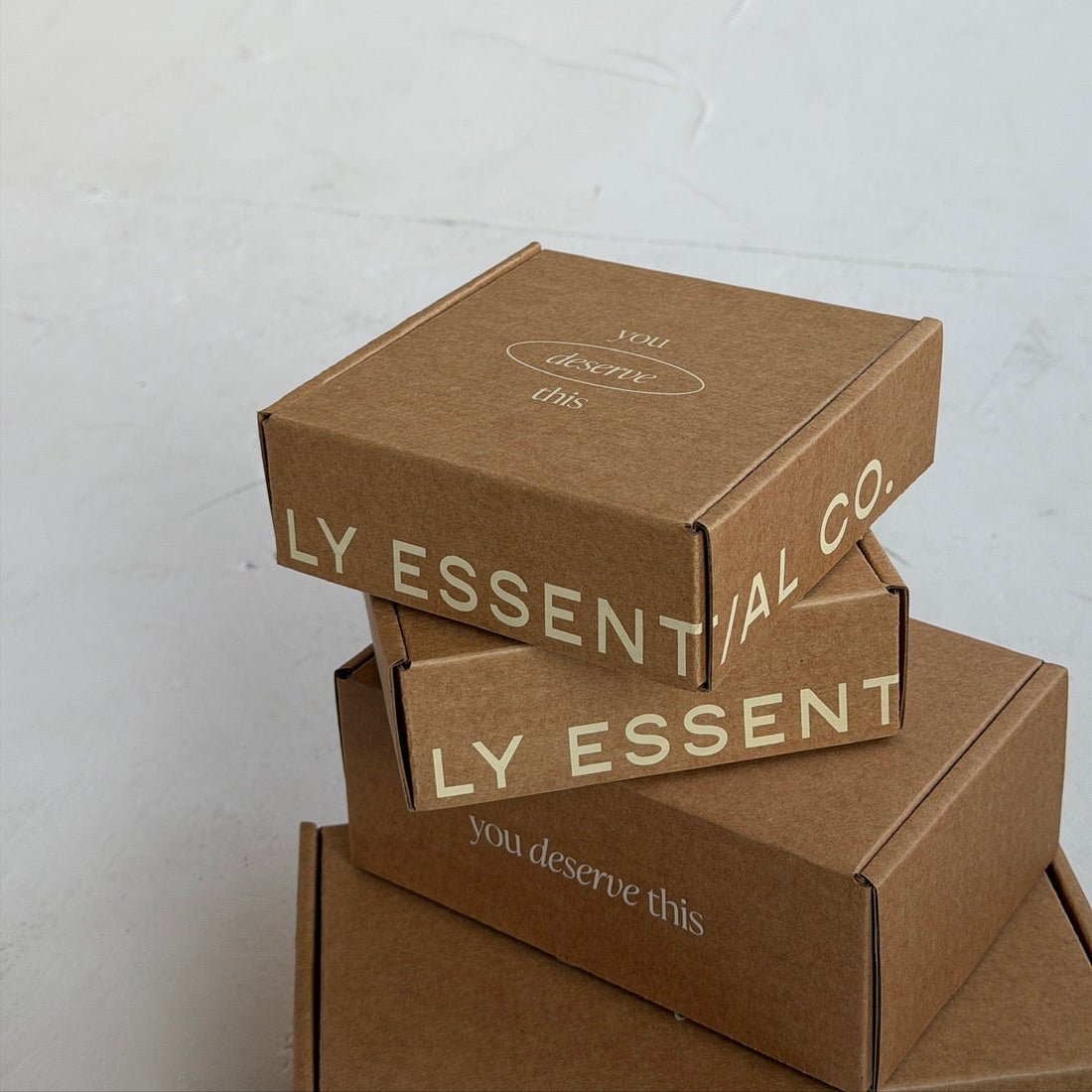Three stacked brown kraft cardboard boxes with beige printed text reading 'LY ESSENTIAL CO.' and 'you deserve this' on a light gray wall.