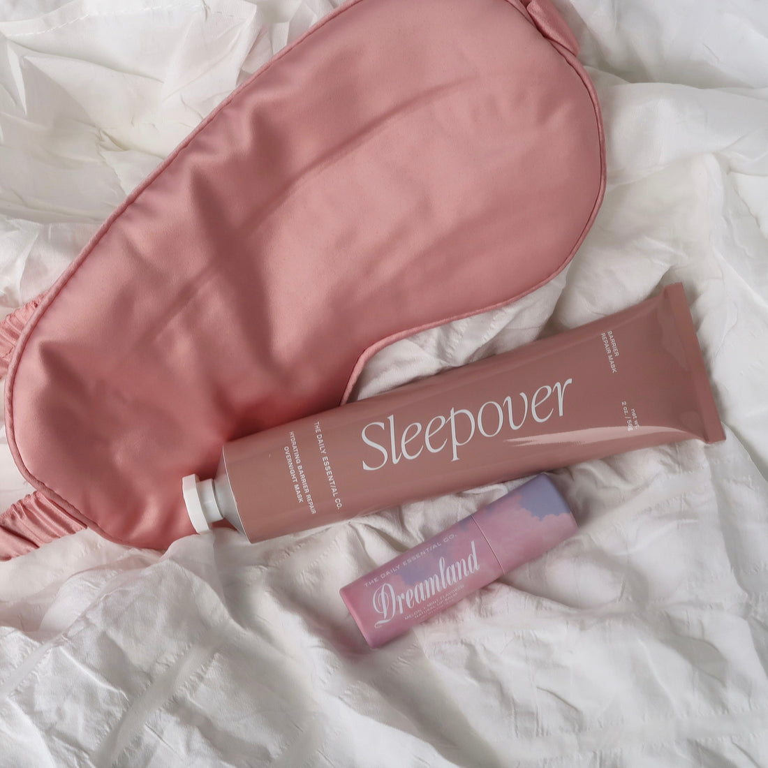 A pink satin sleep mask and a matching pink tube labeled 'Sleepover' arranged on rumpled white bedding, with a small Dreamlight box below.