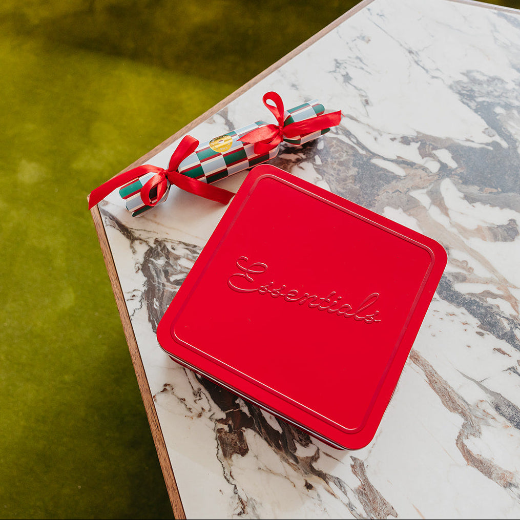 A red square tin with the brand name 'Essentials' embossed on the lid, tied with a red ribbon, resting on a white and gray marble-patterned table next to a green outdoor surface.