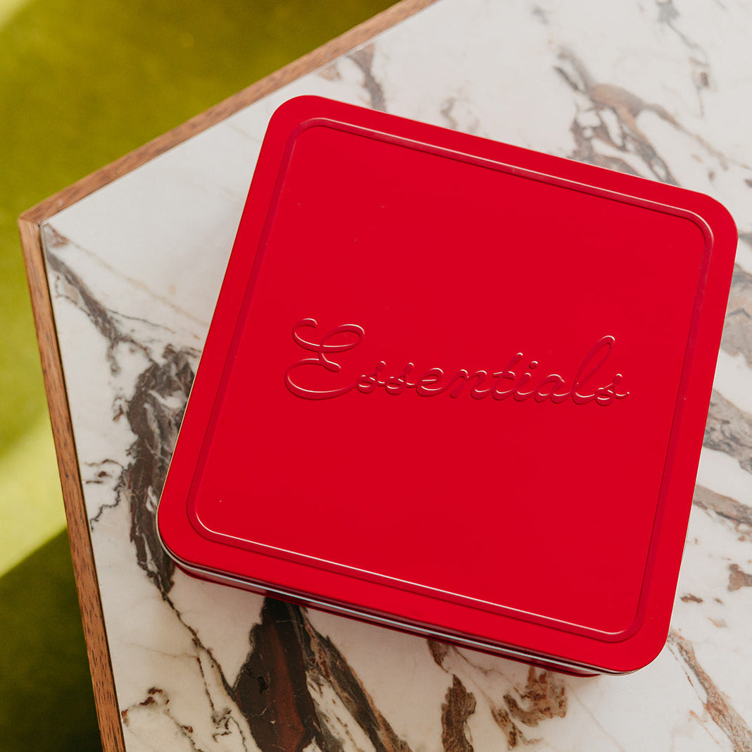 A square red tin with rounded corners and the word 'Essentials' embossed on the lid, placed on a white marble-patterned surface with a bright yellow-green background.
